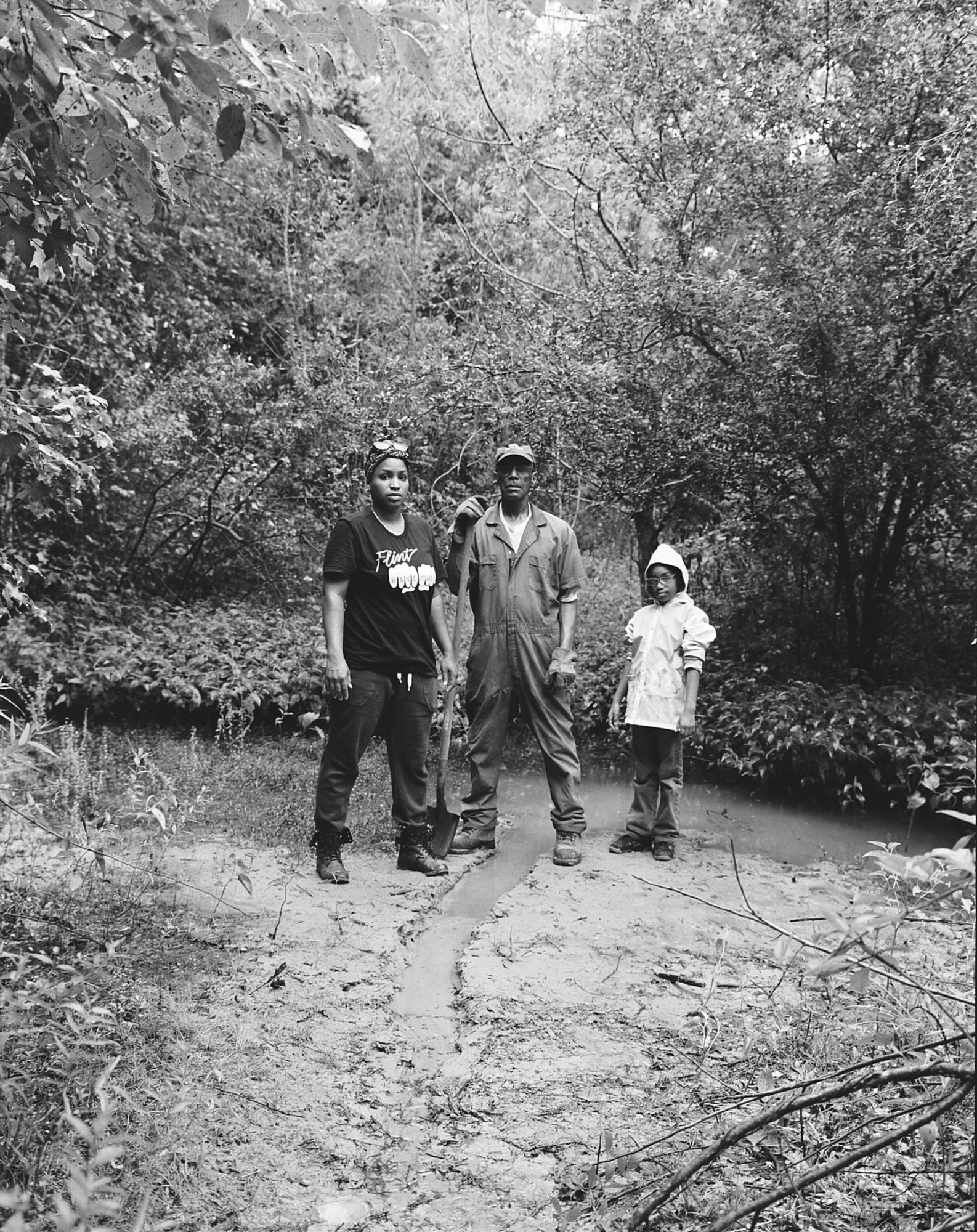 A black and white family portrait containing three generations; grandfather, daughter, grandaughter stand almost defiantly in center frame among trees and next to a freshwater spring