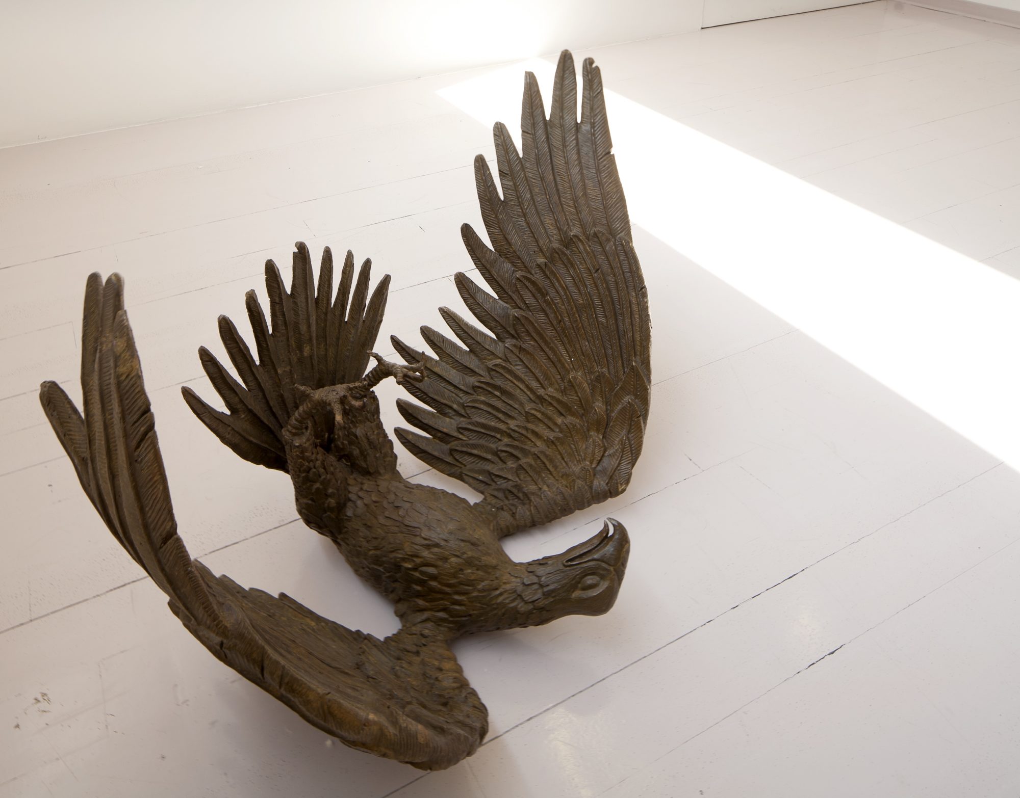 A bronze eagle statue laying on its back with upward sweeping wings and talons, set against a white wooden floor