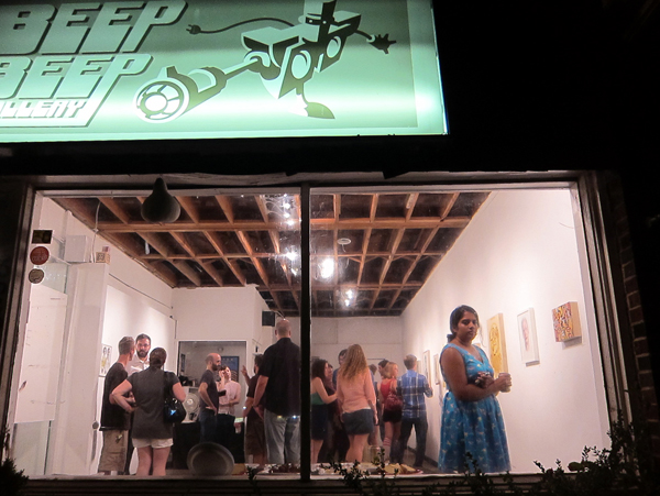 A photo, taken from outside a window, of individuals standing in Beep Beep Gallery looking at art.