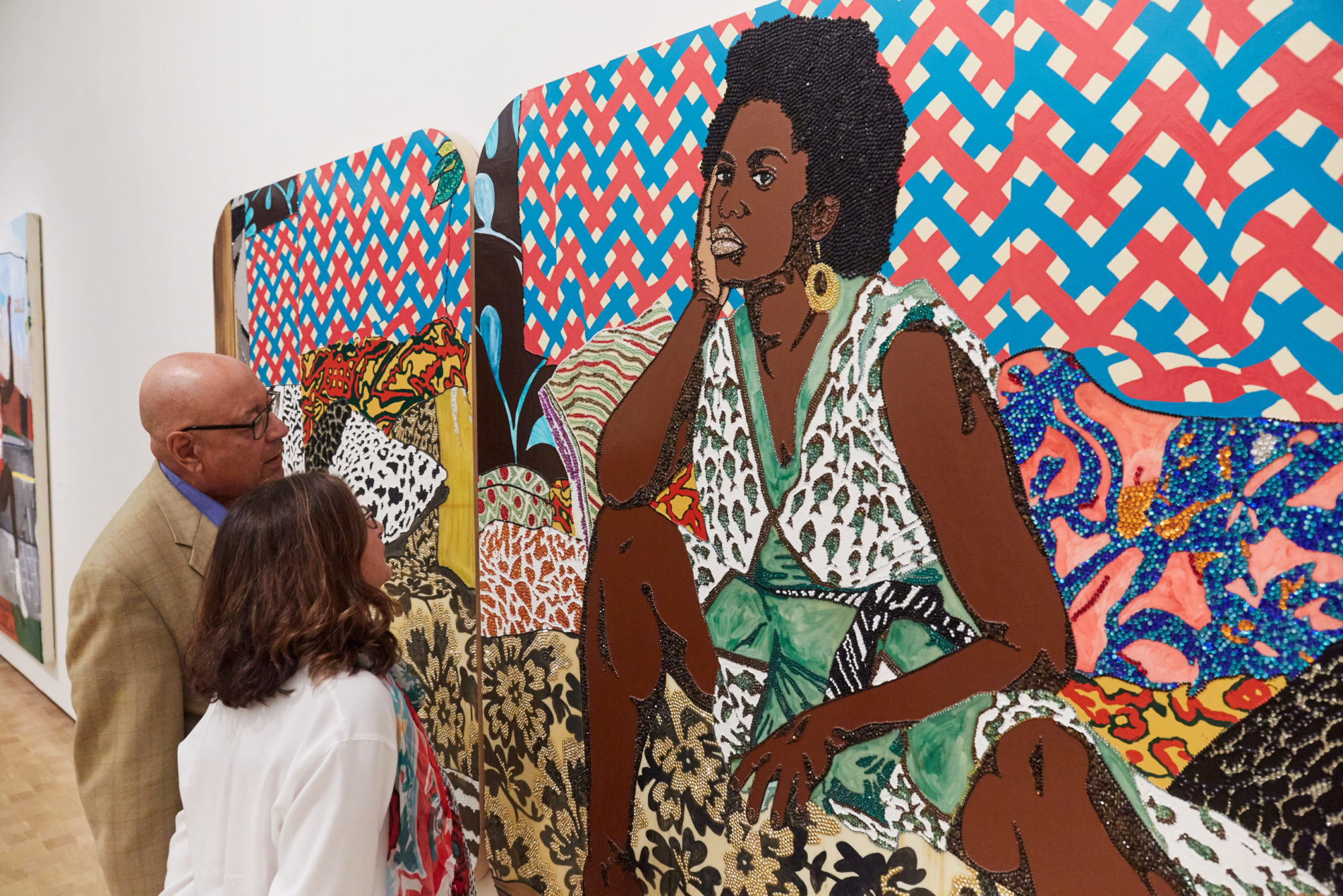 Two museum-goers examine up close a large-scale portrait of an African American woman adorned and sitting amongst varying patterned materials