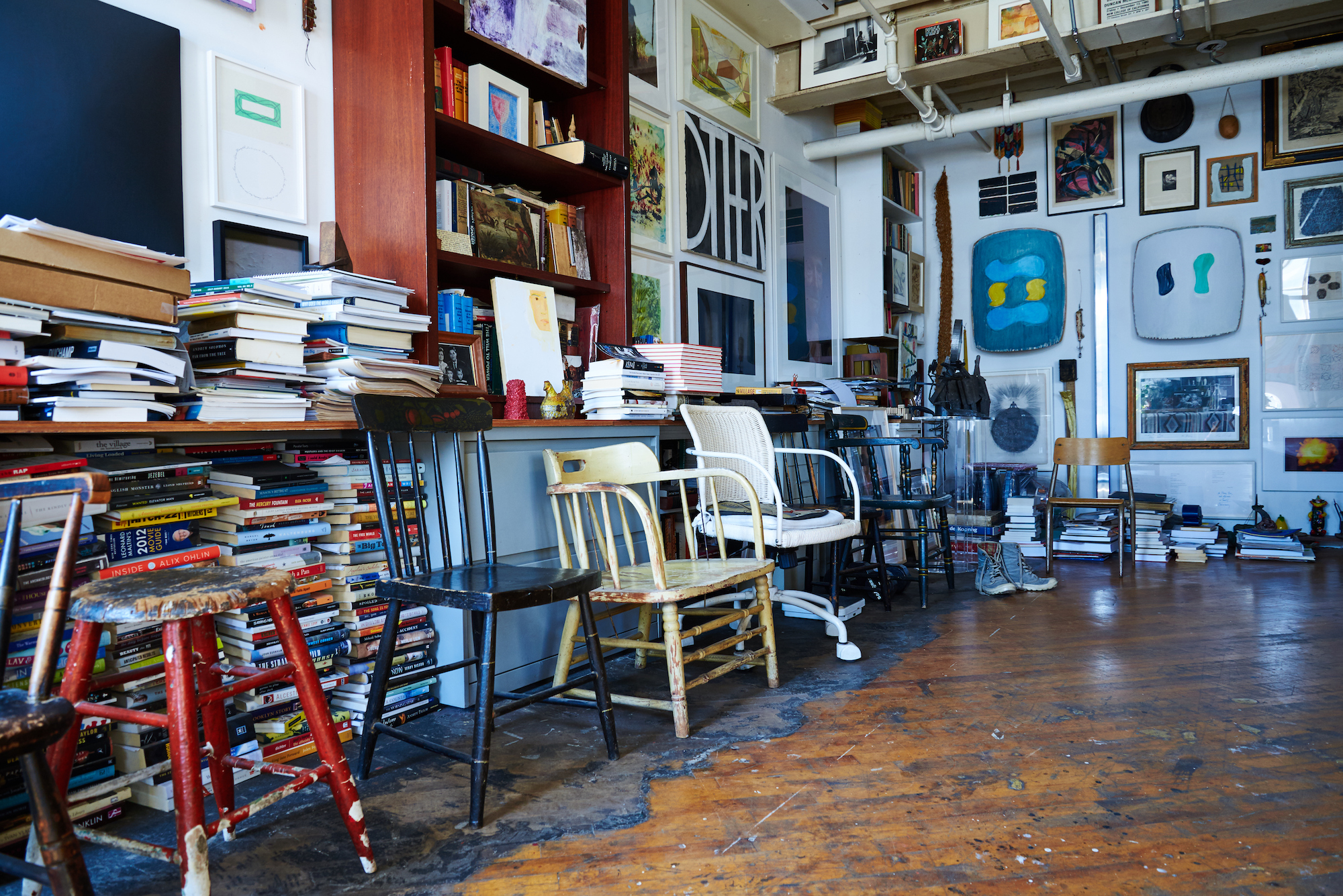 A room with stacks of books, artwork on the walls, and chairs lined up against a wall.