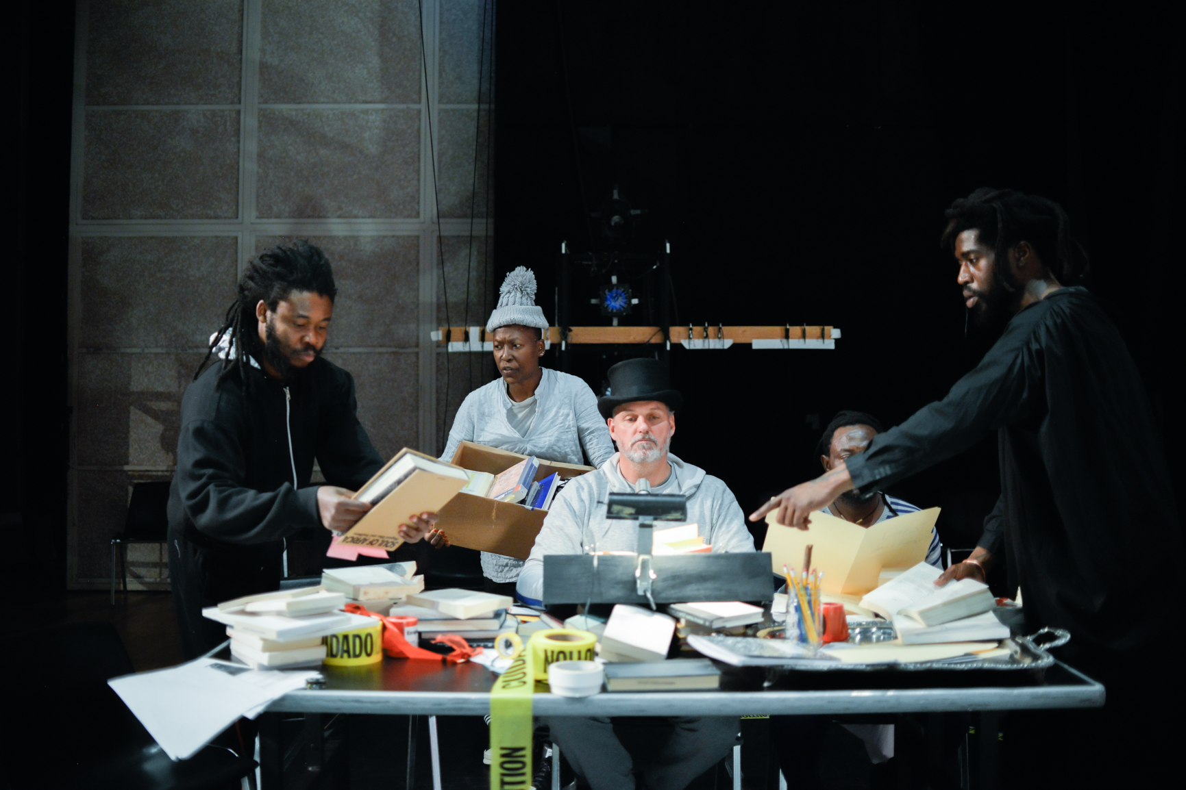 a white man in a top hat and four black people interact with a cluttered table