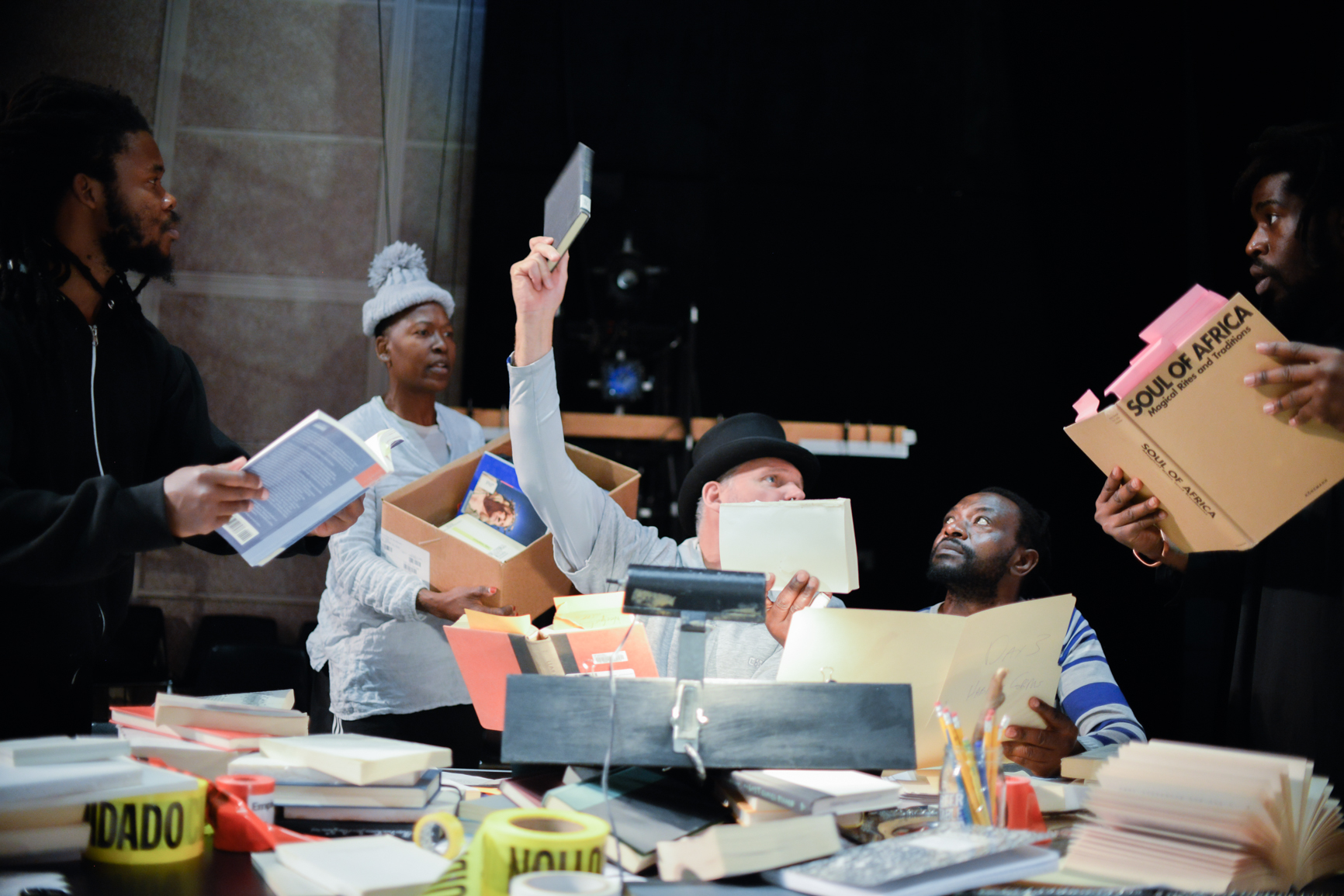 a white man in a top hat and four black people interact with a cluttered table