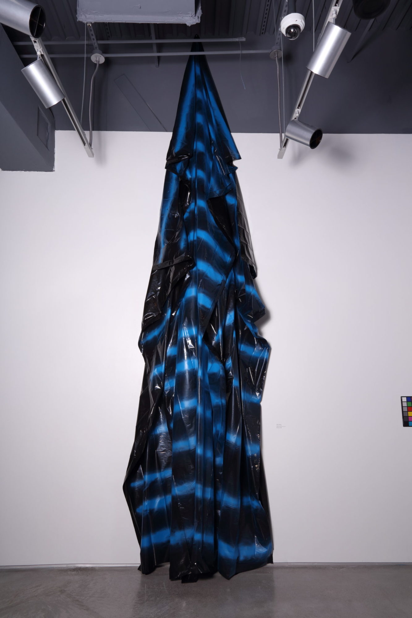 A large blue and black striped form rises vertically against a white wall
