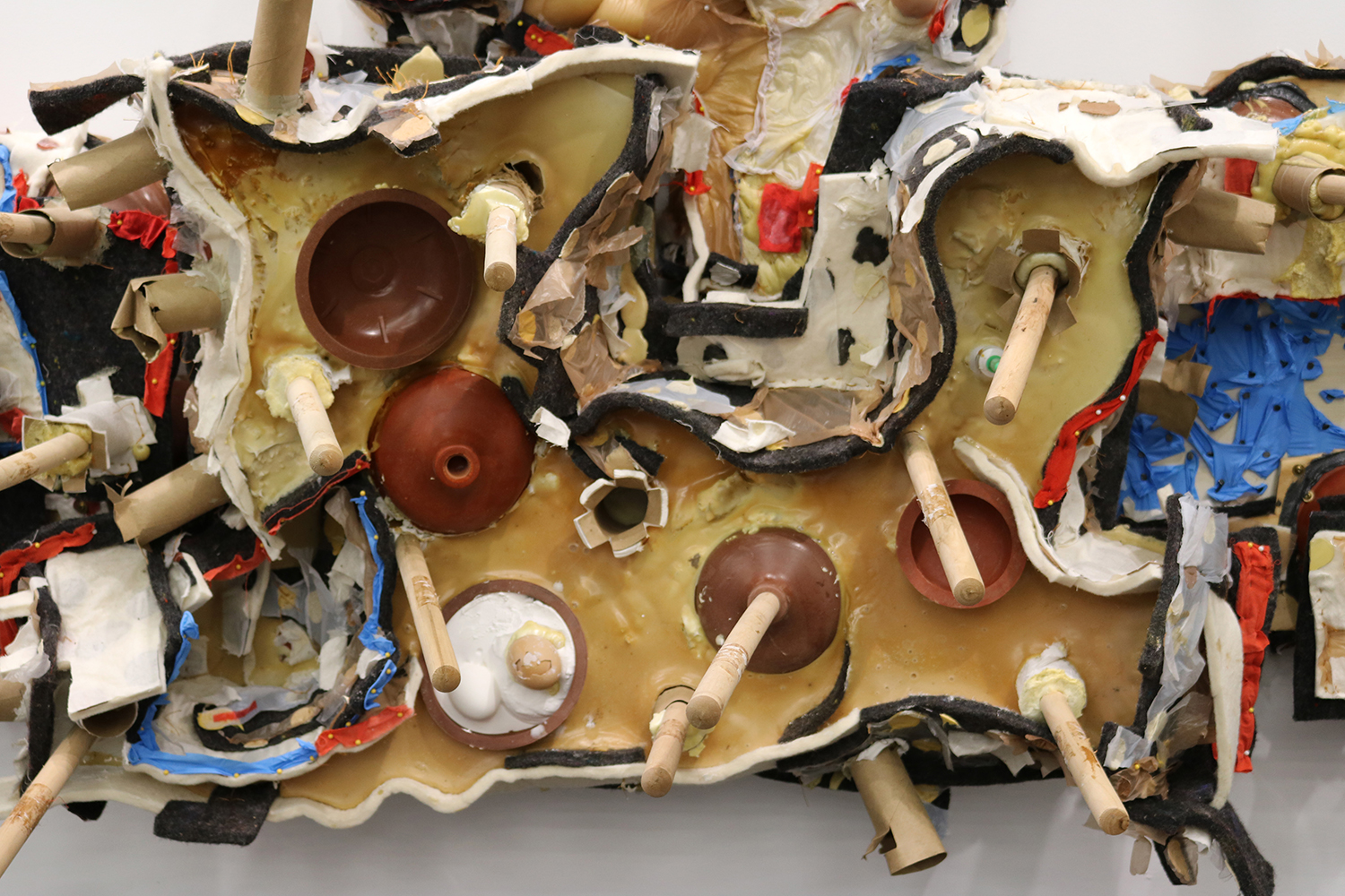 A detail shot of a large, densely packed wall sculpture. This imade shows a funky organic shape containing toilet plungers emerging from and sinking into a pool of latex rubber. One of the toiler plunger heads is filled with plaster and different colored eggshells. Spent toilet paper rolls create decorative orifices in the latex rubber mass, and hints of a harsh electric blue color are created with collaged nitrile medical gloves.
