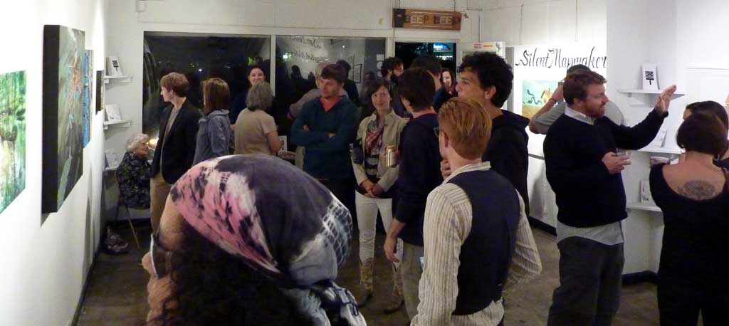 A photo of a gallery. Several individuals stand and look at the art.