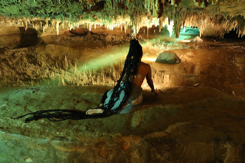 in the interior of a green-lit cave, a woman lounging, her back to us and her hair in long black braids