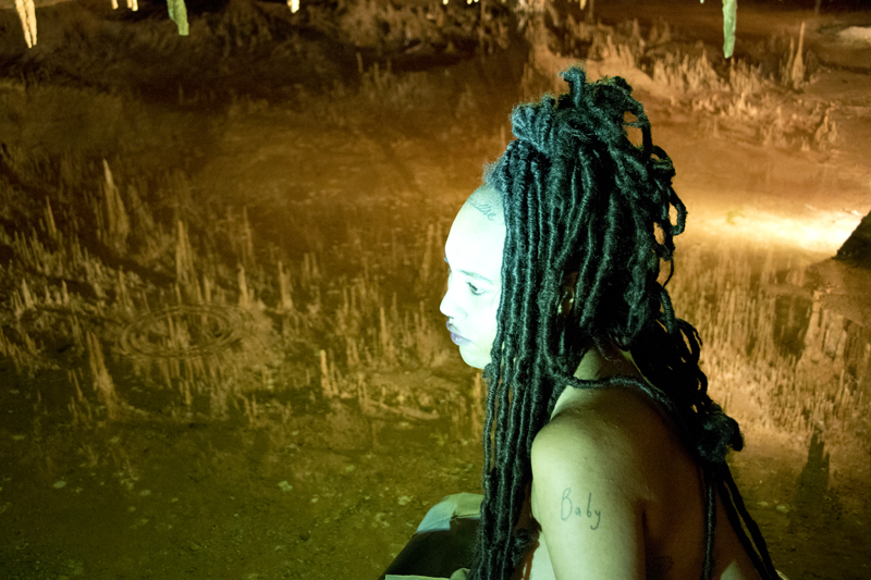 A photograph in a cave; in the background, we see stalactites and in the foreground a seated portrait of a woman with long black coiled hair