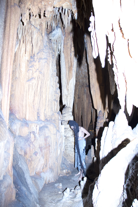 A woman stands in a cave among rock formations; she has long coiled black hair and is wearing only a blue-grey skirt