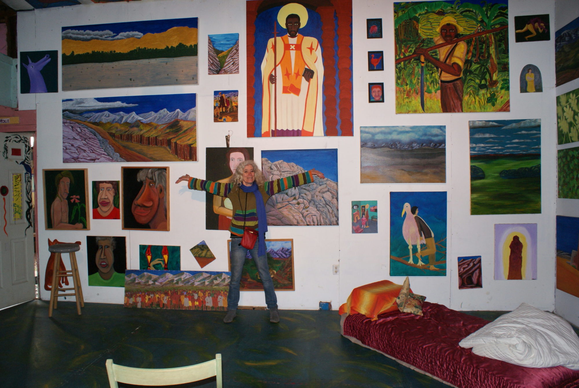 A photograph of Cafe Bizzozos. The back wall is covered in colorful art — close to 20 paintings. One individual holds their arms out and smiles at the camera. A red velvet mattress is on the floor in one corner.