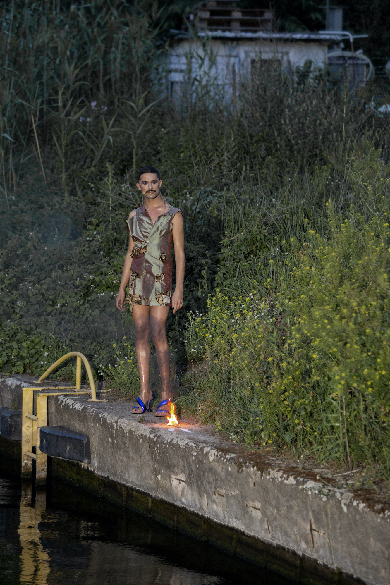 a masculine person with a mustache stands amid wildflowers while wearing a short shiny garment and high heels