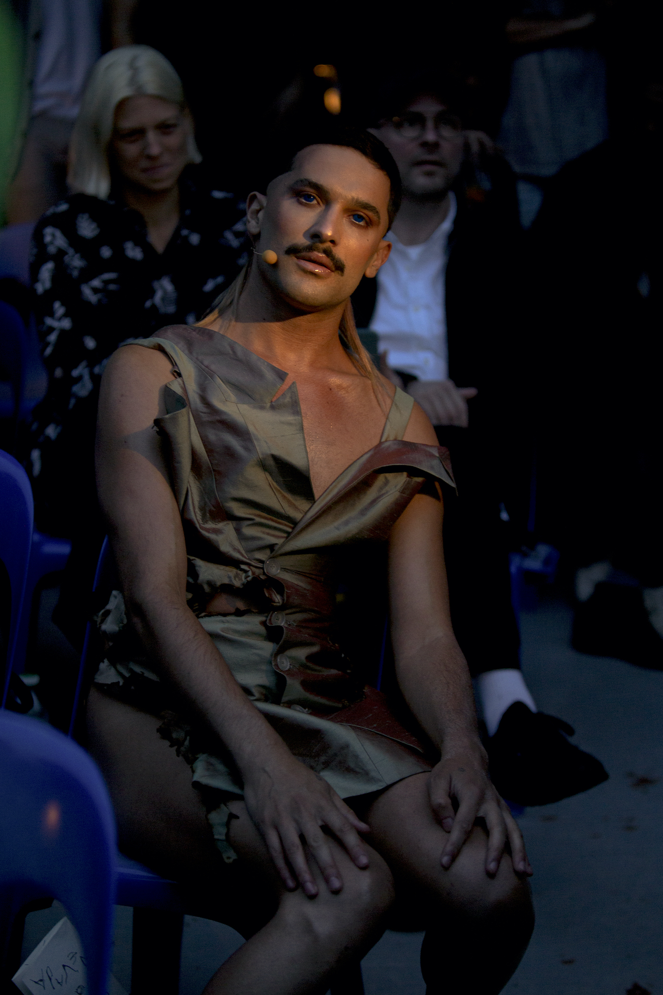 a masculine person with a mustache wears a shiny garment and sits