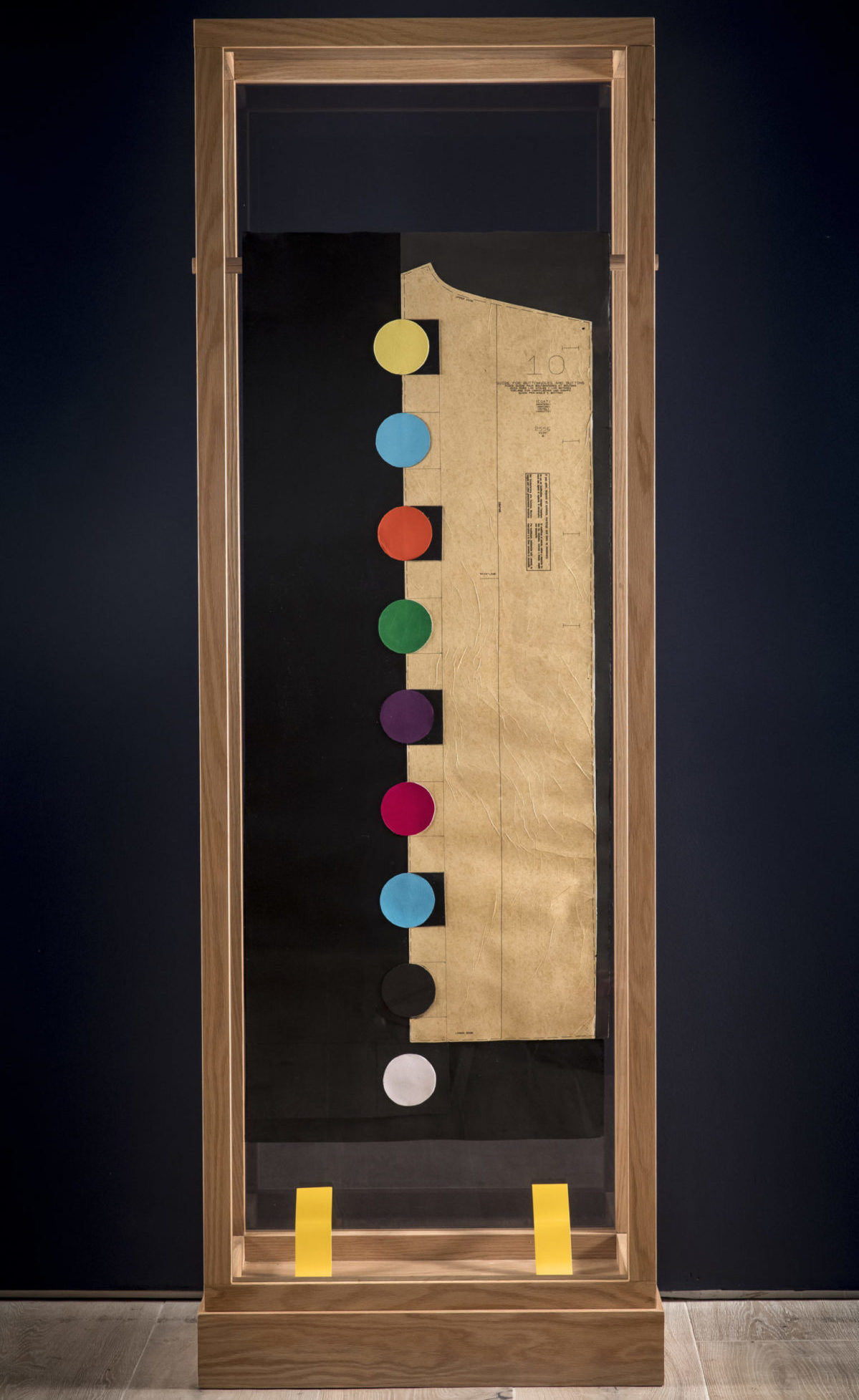 Image of Derrick Adams work, nine multicolored circles in a vertical line bisecting a field of black from measurement paper for a garment