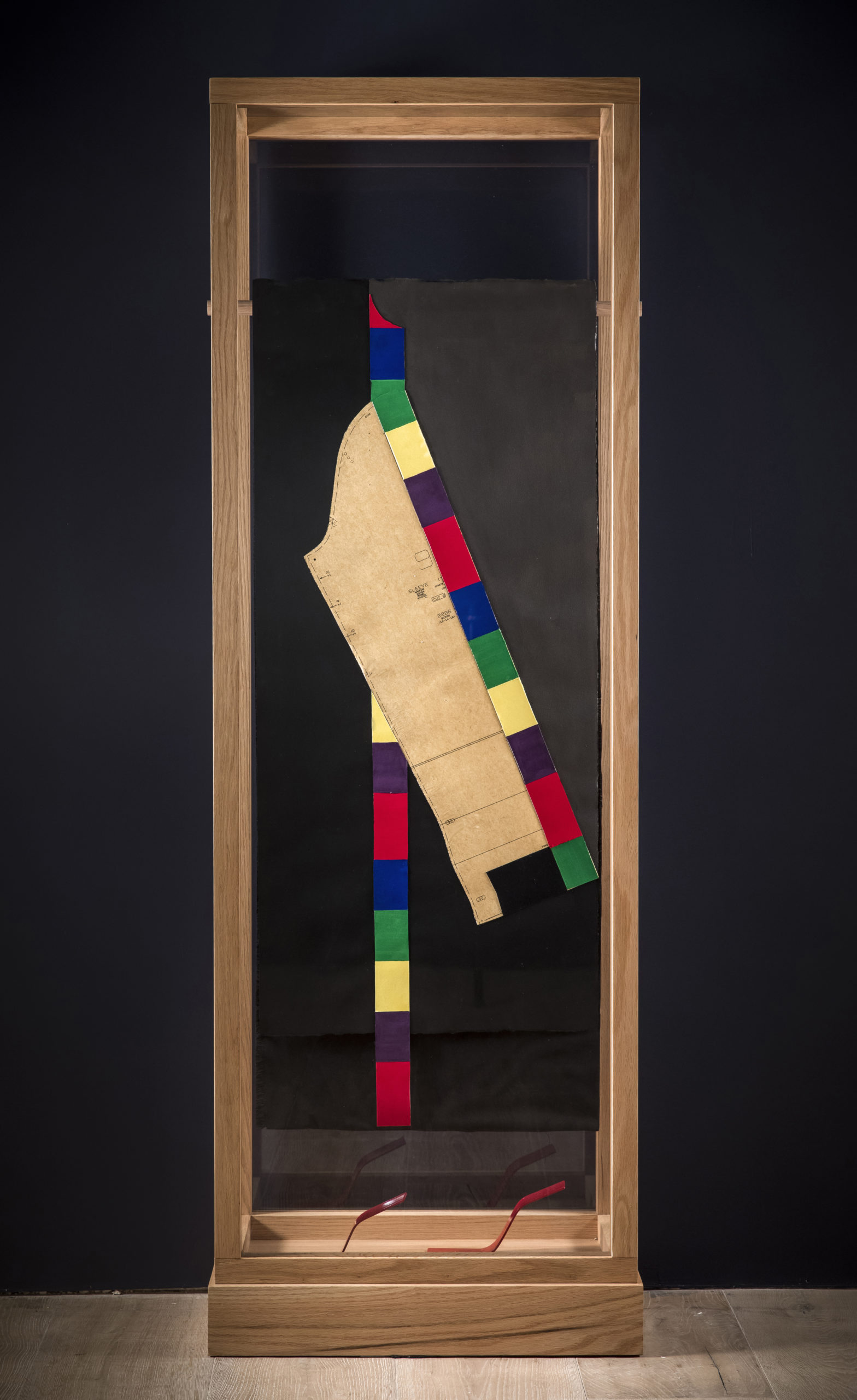 Shot of Adams' piece enclosed in a wooden box with glass sides; inside hangs a black paper with multi-colored stripes running up one side and the measurements for a garment hanging down the middle; two red paper heels rest on the floor of the box