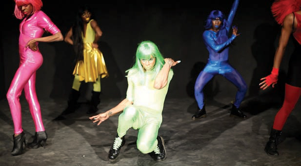five dancers strike poses, each in a different monochrome look including a wig