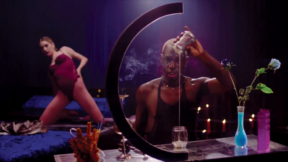 a video still from Hush Bb, showing singer and performer Le1f pouring wax from a candle into a glass. A dancer wearing a pink leotard dances on a bed in the backgroud.