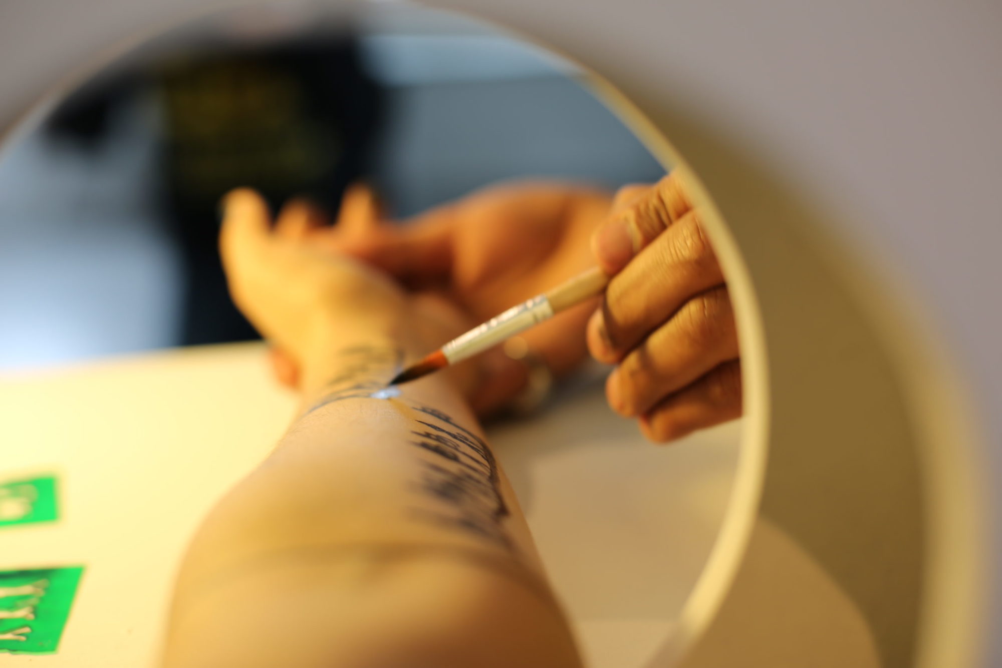 photograph of the artist painting solid black figures on a forearm which is stretched through a hole in the wall