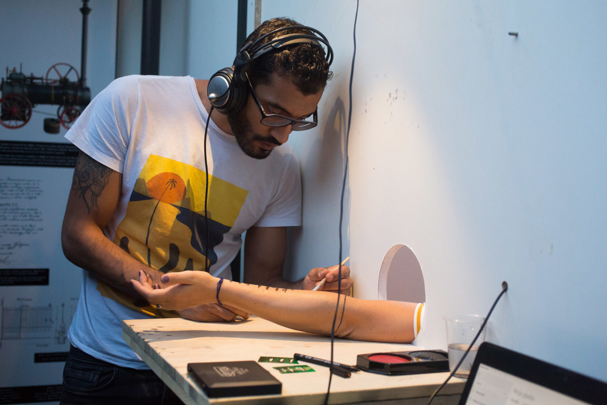 photograph of the artist painting solid black figures on a forearm which is stretched through a hole in the wall