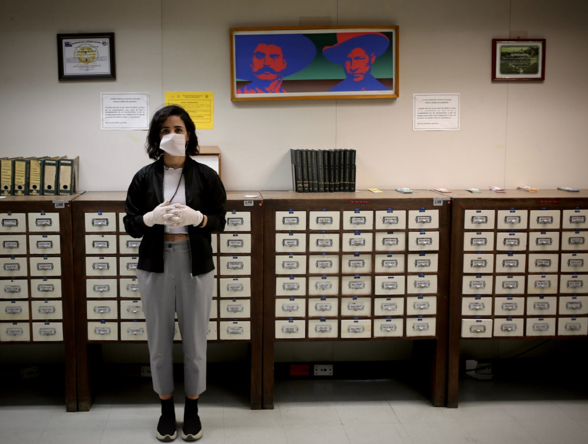 a woman stands in front of a group of labeled drawers; she is to left of frame, feet and hands together, a medical mask across her mouth; she stares directly into the camera