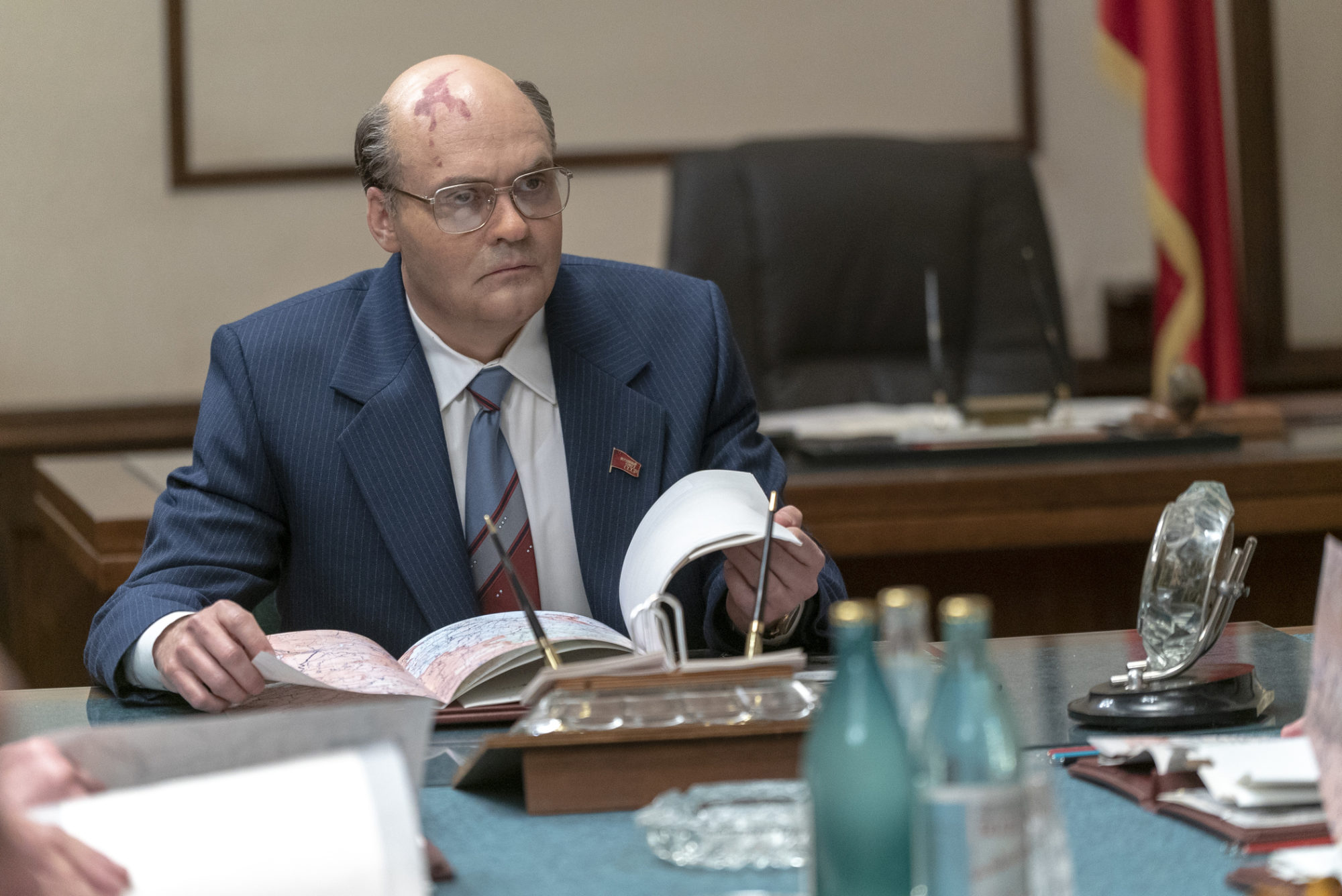 David Dencik wears a suit and sits at a desk with a map and open file in his hands