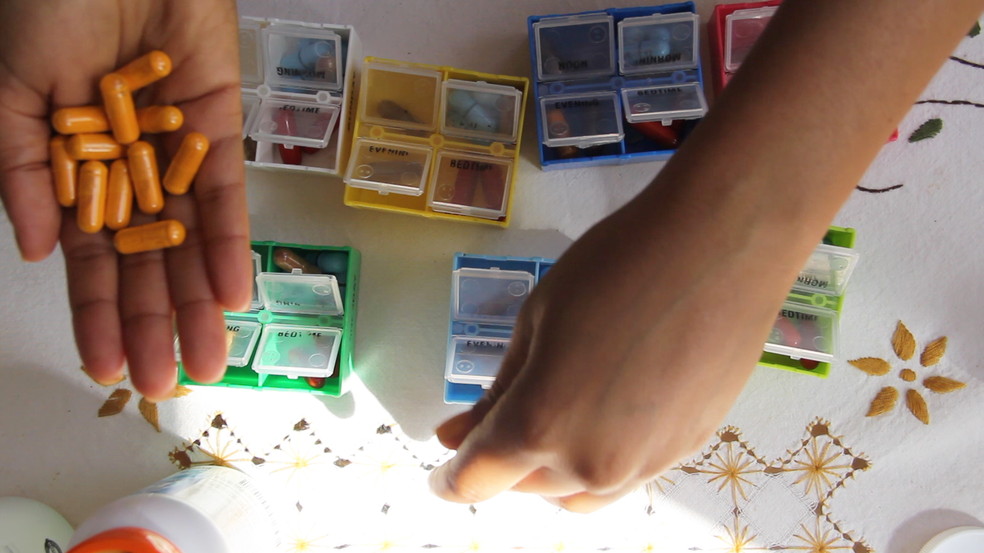 Two hands sort pills into seven brightly colored pill boxes on top of an embroidered cloth.