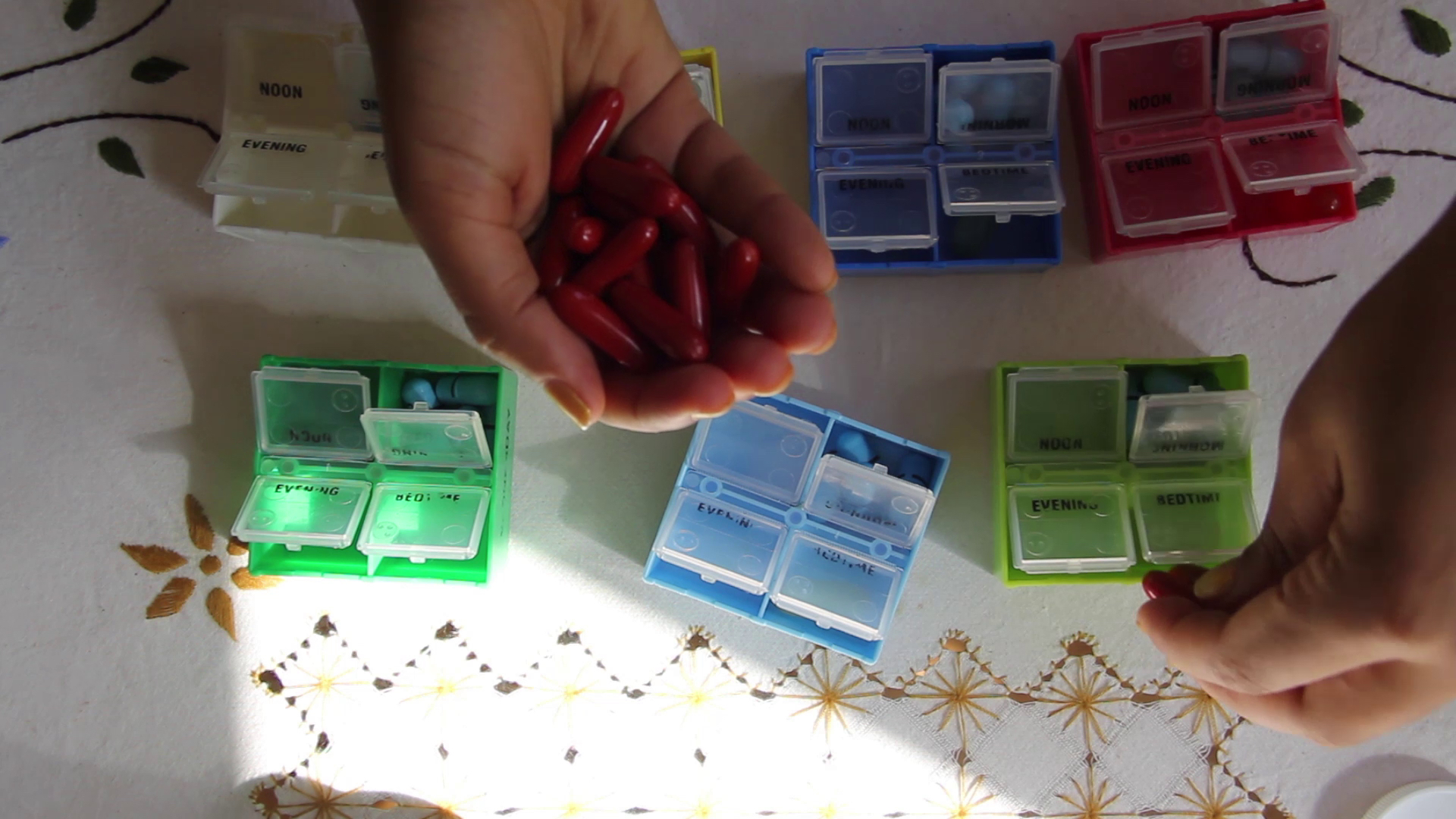 Two hands sort pills into seven brightly colored pill boxes on top of an embroidered cloth.