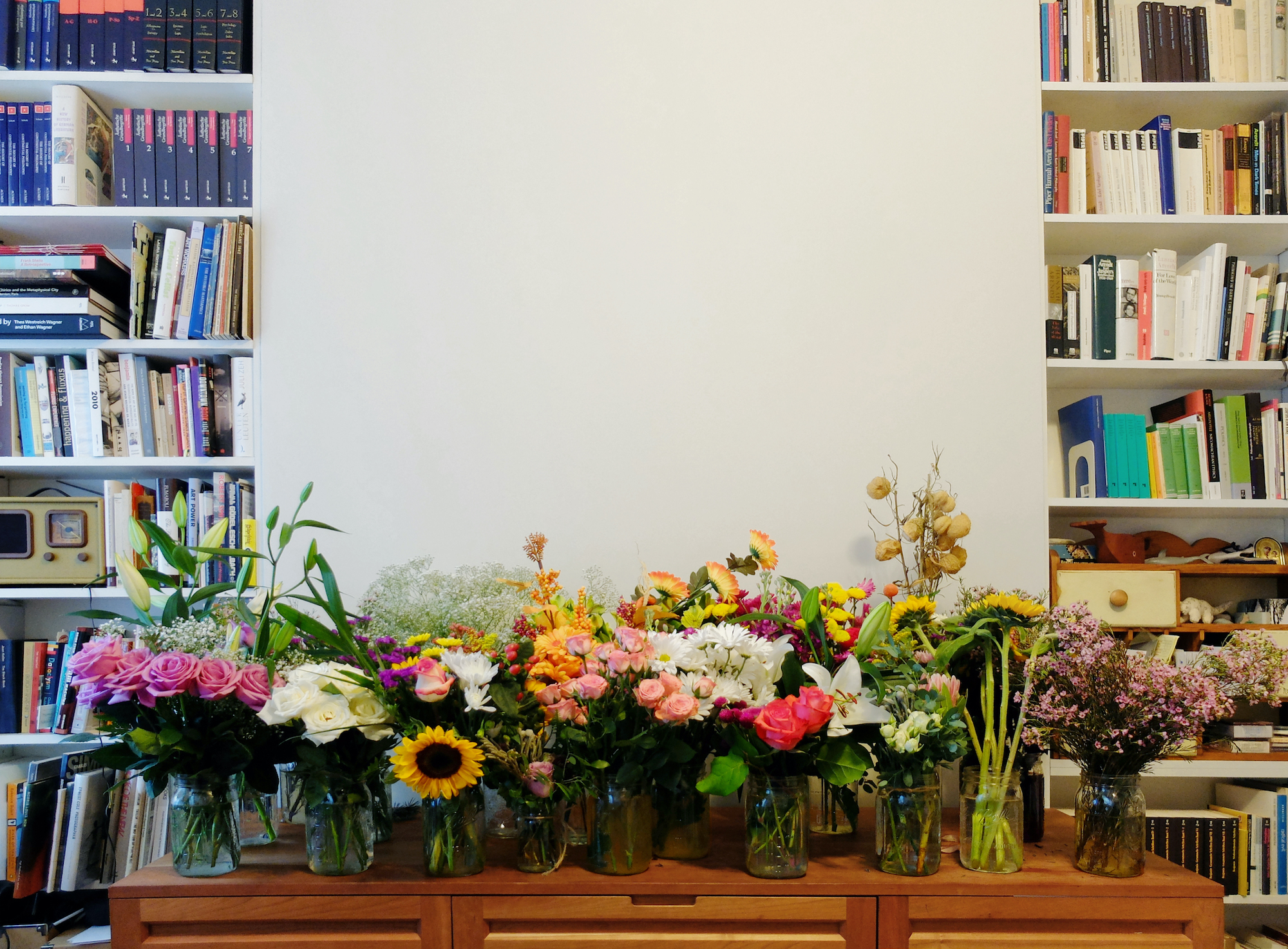 An assortment of flower bouquets in jars and vases against a tan wall.