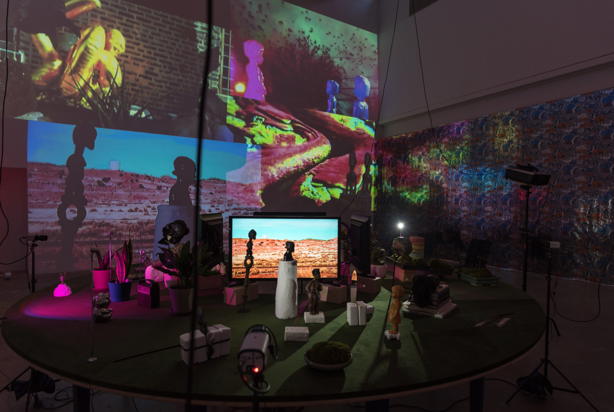 various objects are located on a round table in the center of a dark room. video projections of the items on the table are displayed wall