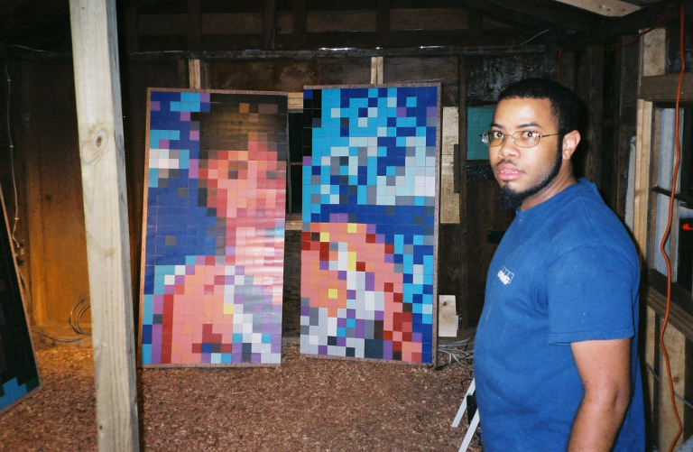 An image of an individual standing in a shed in a blue T-shirt standing next to a piece of artwork that resembles the individual but in pixelated form.