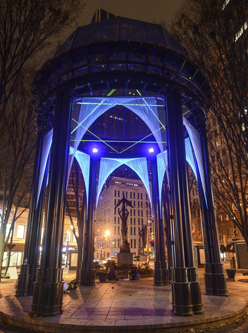 Image is of a gazebo (Woodruff ParK) which has been strung with neon black-lit blue strings and whose ceiling boasts a neon black-lit green Star of David by Megan Mosholder