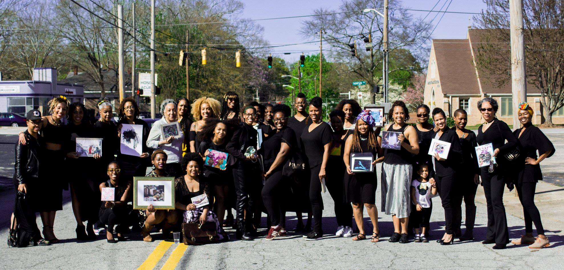 A photograph of around 30 individuals standing in the middle of a road dressed in black and holding pieces of artwork.