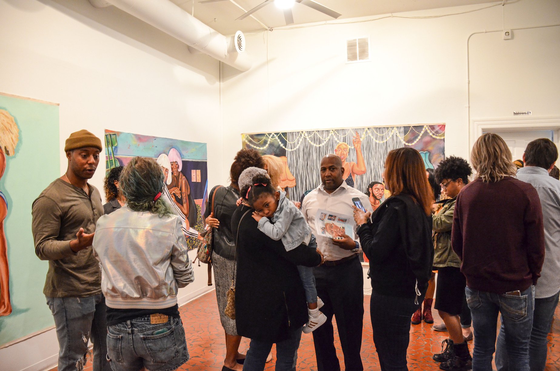 A photograph of a gallery. Around 10 people stand in the middle of the room making conversation and looking at the art on the wall.