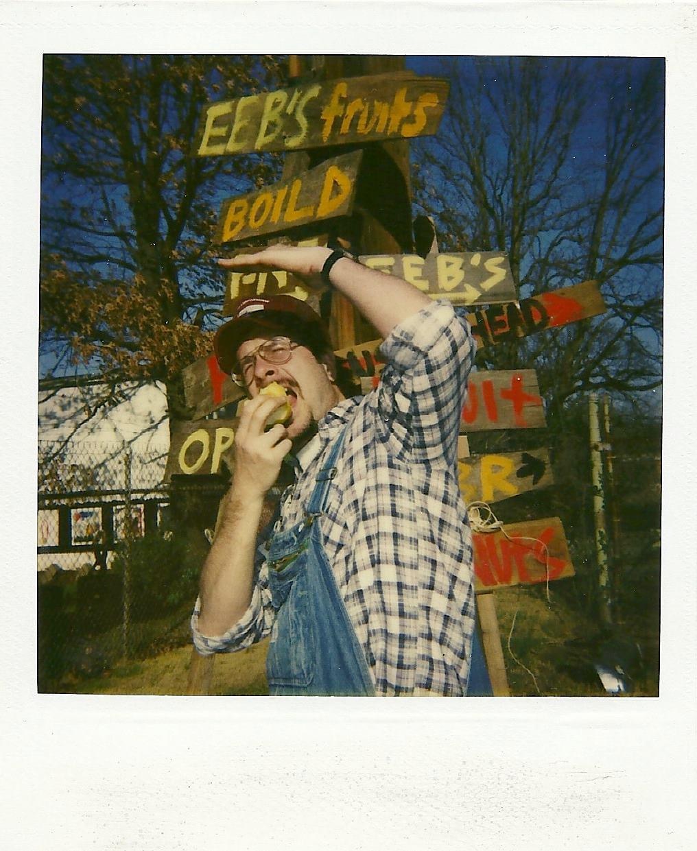A photograph of an individual in a plaid shirt and denim overalls eating an apple standing in front of a set of signs — the top sign reads 