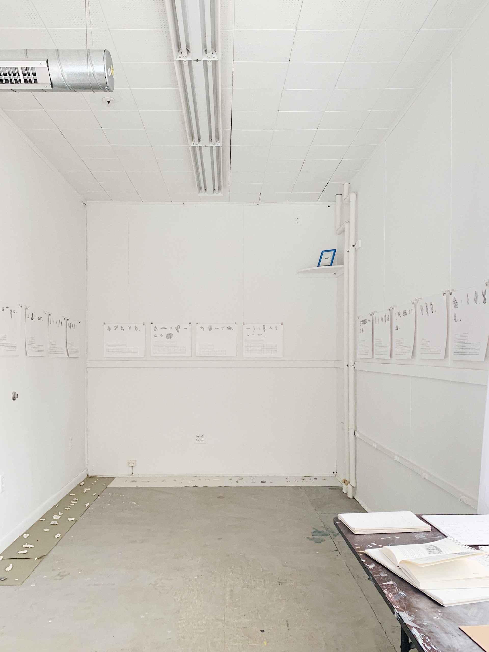 The inside of a white studio with concrete floor, a work desk in the bottom right corner with open books on it, and papers with drawings hanging up on the walls.