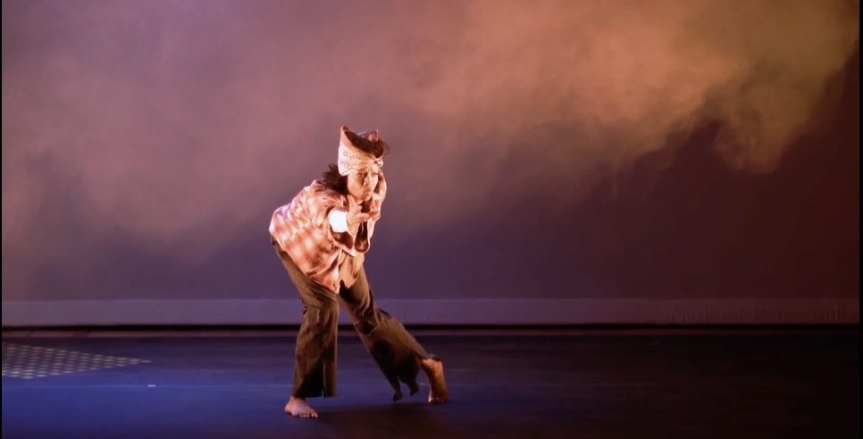 Person (Gesel Mason) standing on a stage barefoot wearing brown pants, a rust flannel shirt and a brown bandana around their head, bent forward with their right arm outstretched.