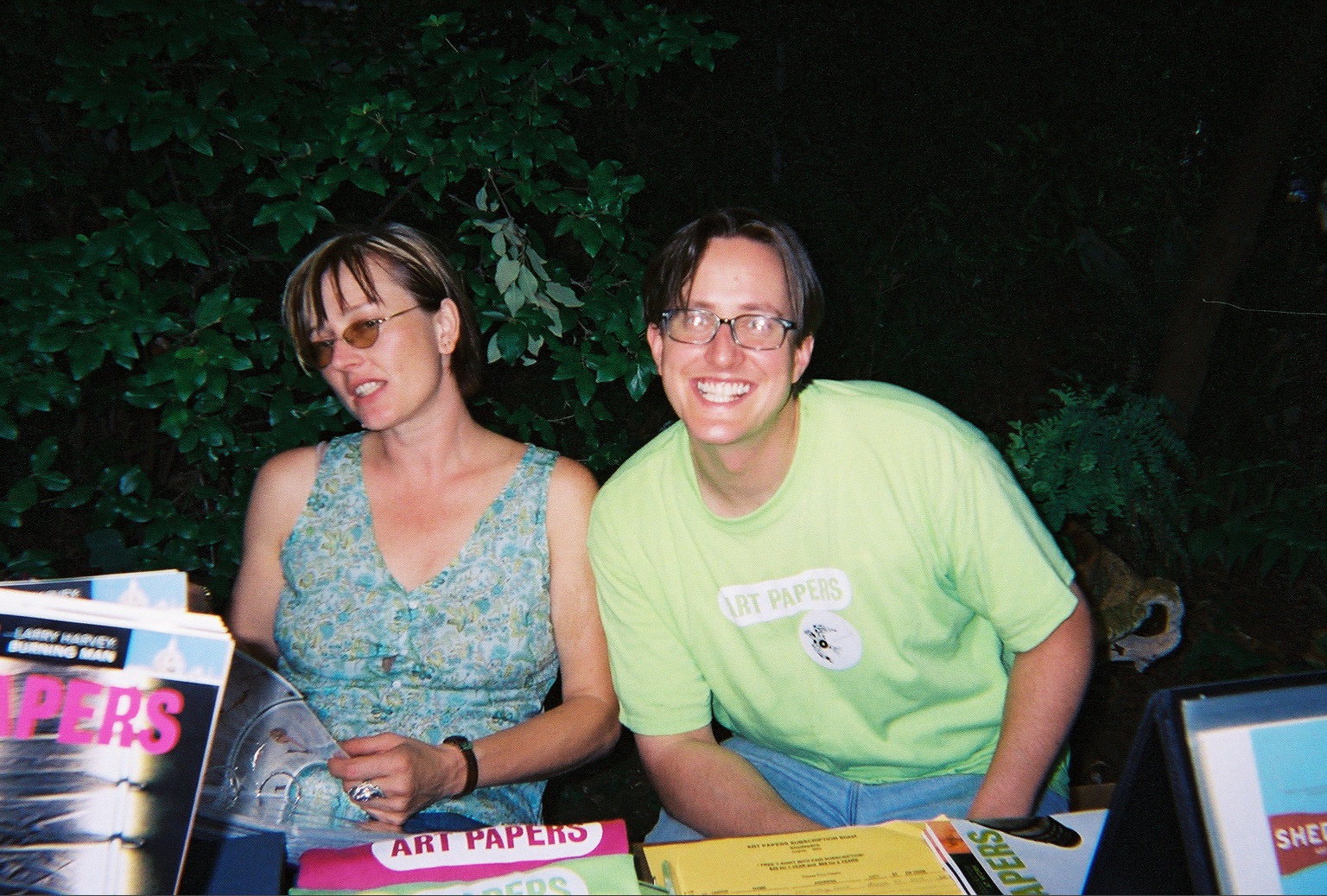 An image of two individuals smiling and surrounded by Art Papers issues and T-shirts. Dark leaves are in the background.