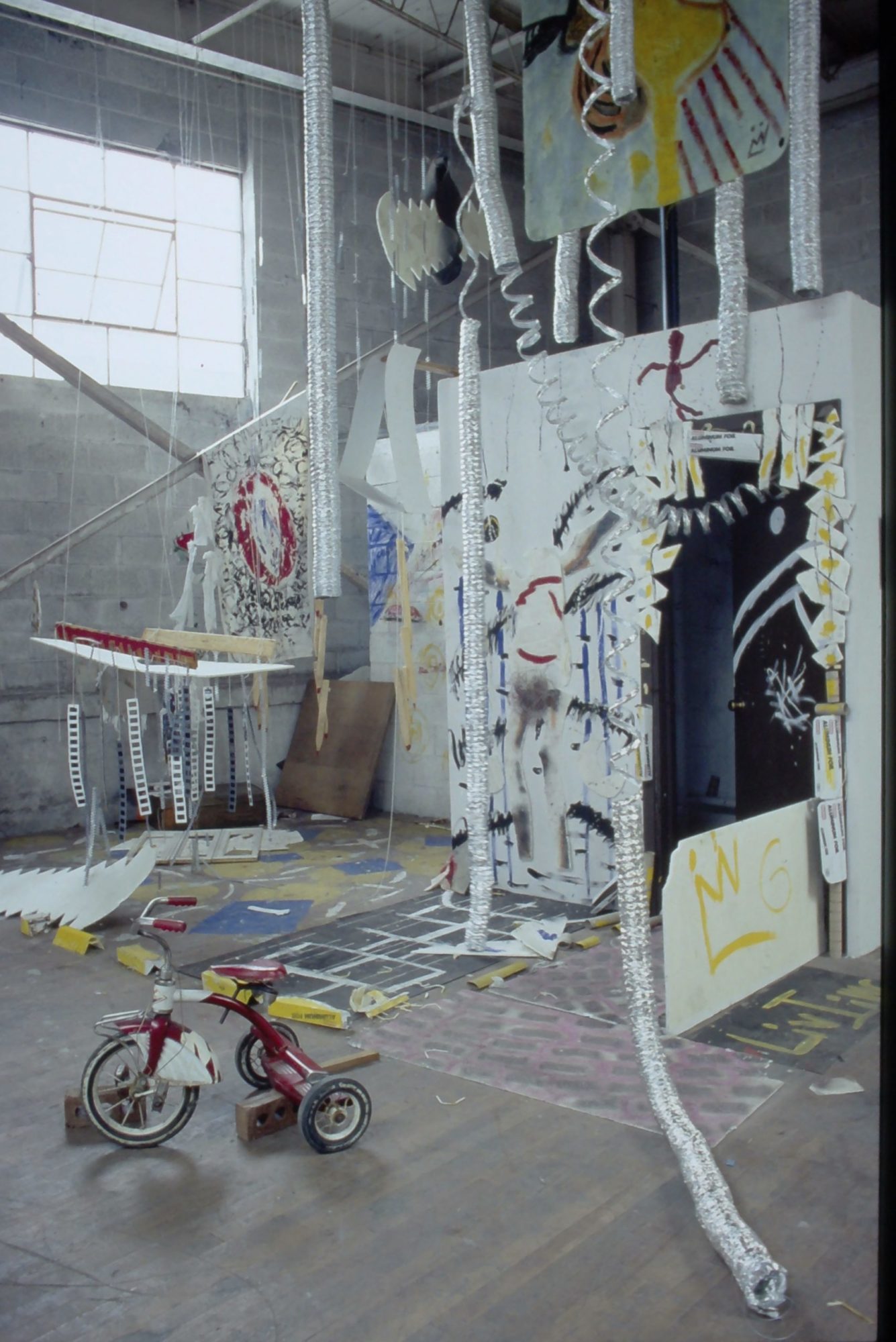 A photograhp of an art installation, which includes several silver hanging tubes and a small tricycle on the ground.