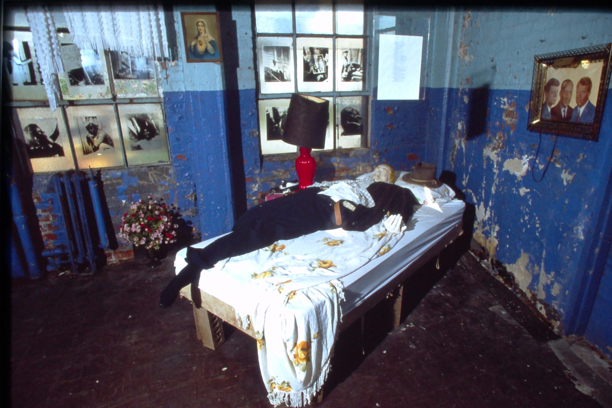 A photograph of someone's room with blue-painted walls — and many chips in the paint. An individual lies motionless on a bed in the center of the room, and many portraits hang on the walls and in the windowpanes.