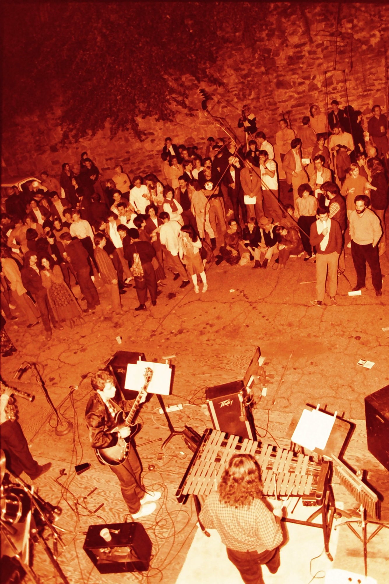 An orange-toned photograph of several musicians performing outside for a standing crowd.