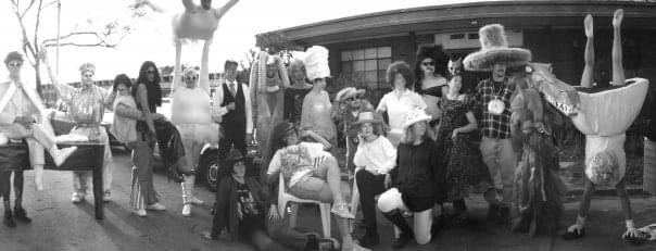 A black and white photograph of several individuals in costume. Some are sitting, others are standing. One is doing a handstand.