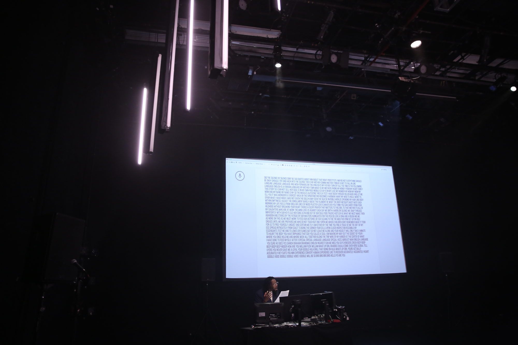 Wide view of person in dark room reading off of pages in front of projection of text