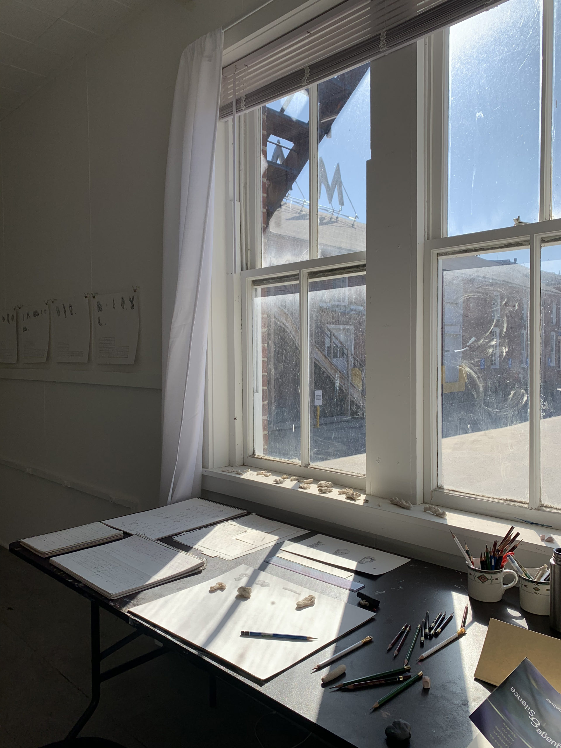 A view of the inside of a studio: a desk with papers and drawing utensils, a window with a view of daylight outside, and three papers hanging on a nearby wall.