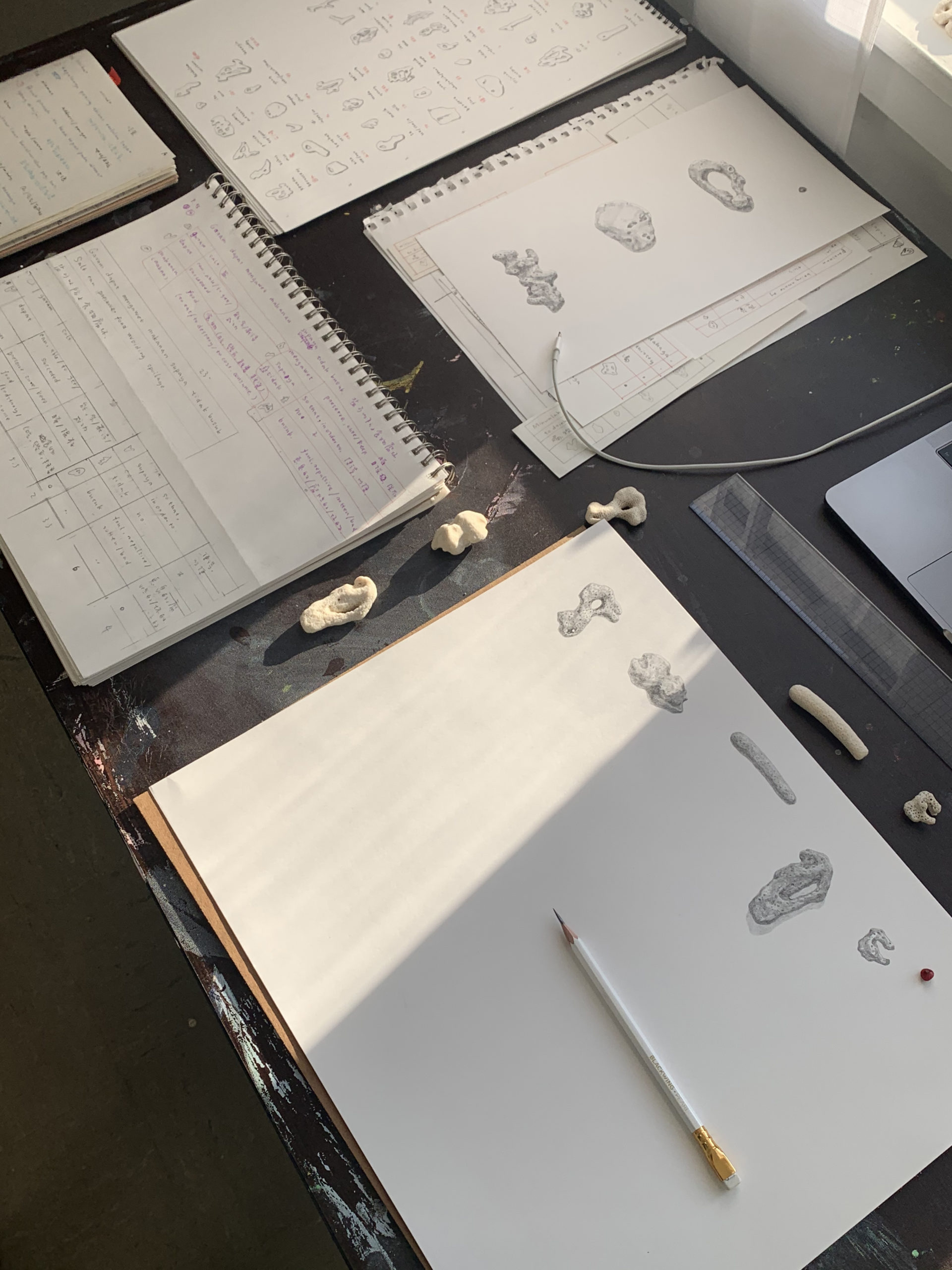 Close-up view of a desk with a sketchbook with drawings of small figures, similar small figures in clay sitting nearby on the desk, and other books and papers beside them.