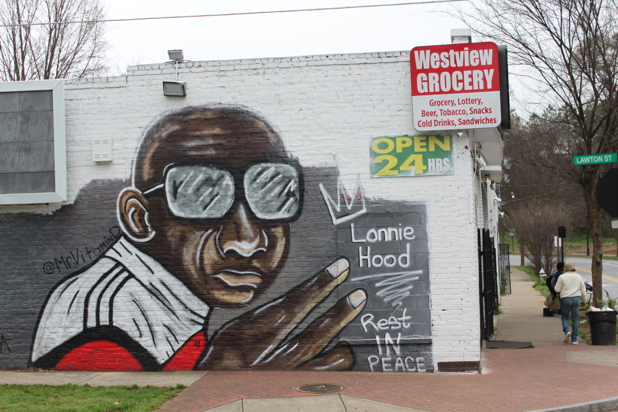 A mural on the side of a grocery store depicts a black person wearing sunglasses and holding aloft a hand. Text next to the mural reads 