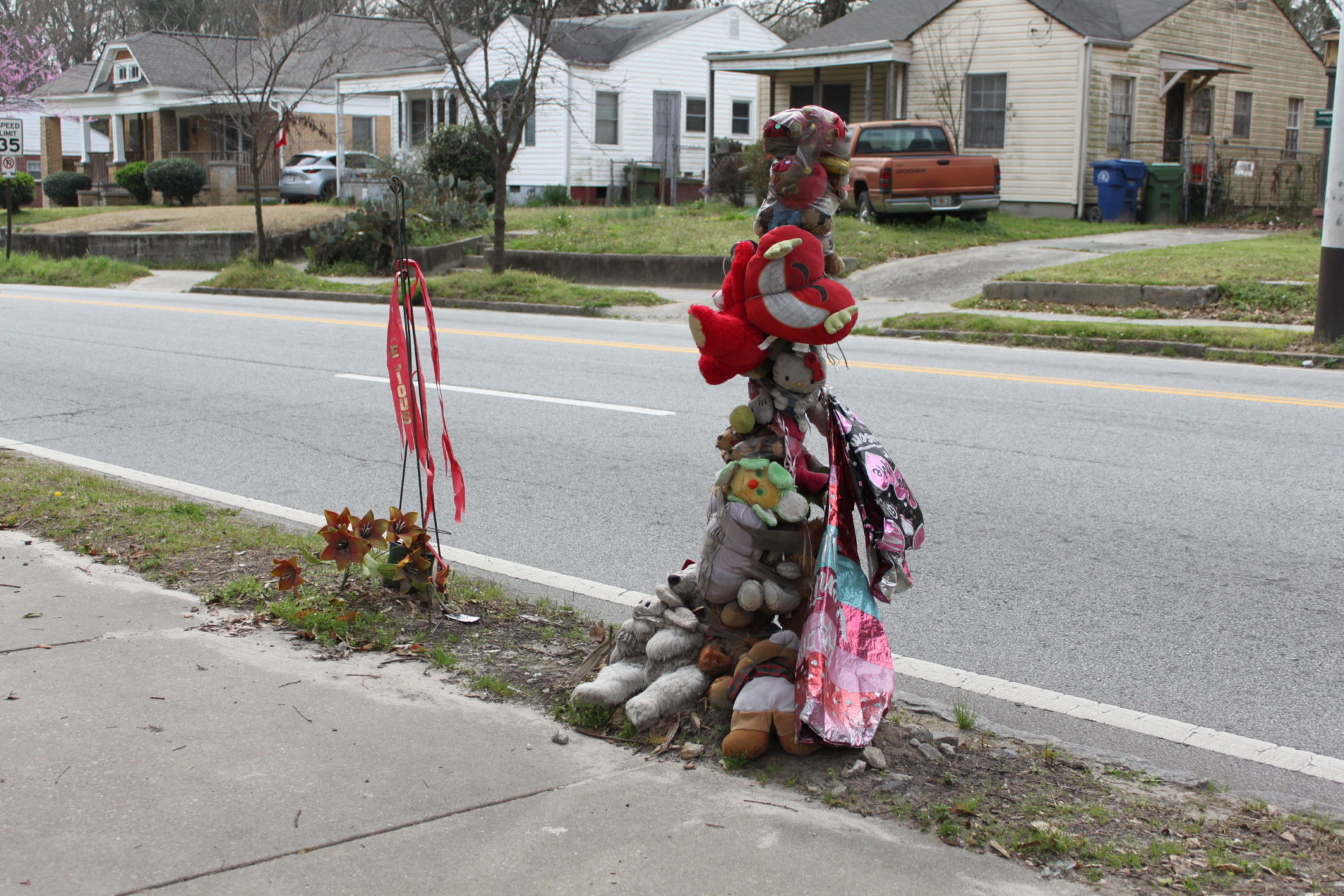 Two poles placed next to a sidewalk are garnished with stuffed animals ribbons and pennants. They are in front of a row of houses