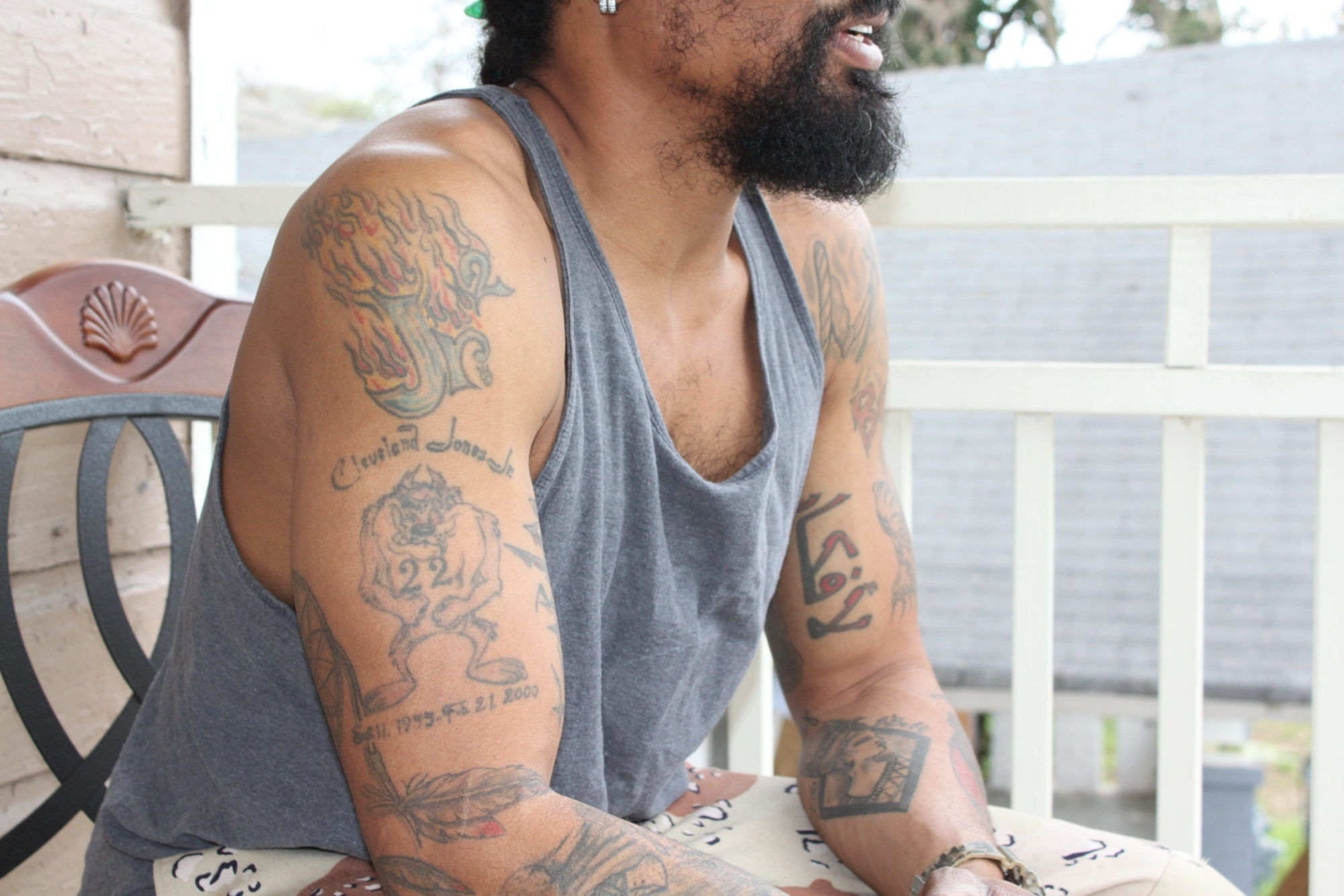 A closeup of a black person's torso as seen from the side shows two arms with tattoos. The person is wearing a light gray tanktop.