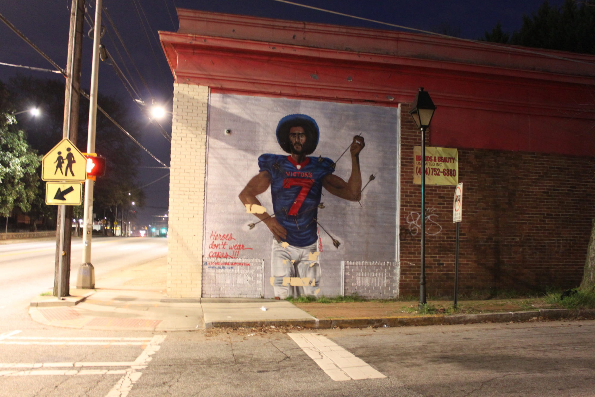 A mural on the side of a red-brick building features Colin Kaepernick wearing his football jersey. He is pierced with arrows and the bottom left corner reads, 