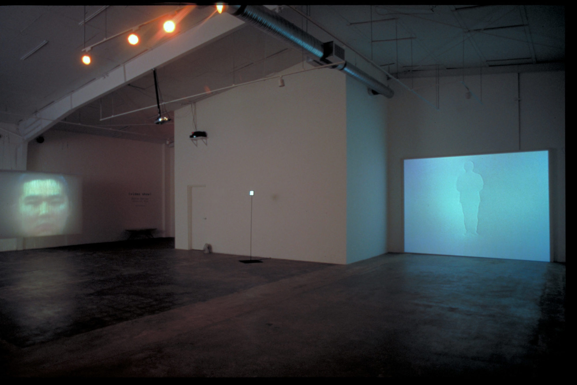 A photograph of an instillation view with white walls and a dark floor. Two images, one of a silhouette and another of a face, are projected on the walls.