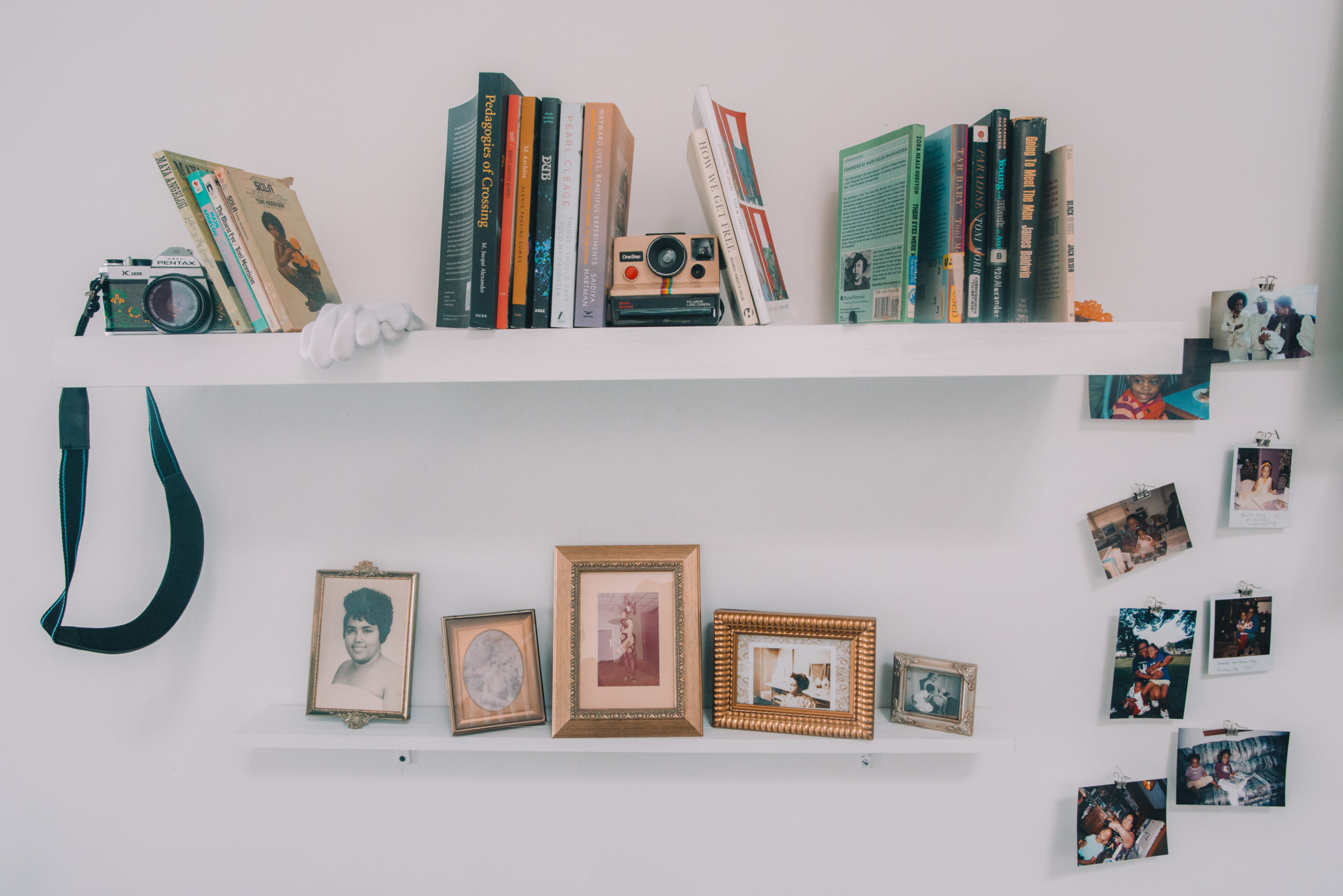 A photo of two white wall bookshelves. The top shelf is longer than the bottom shelf and holds several colorful books and two cameras. The bottom shelf holds five framed photos. To the right of the bookshelves hang eight polaroid photos.