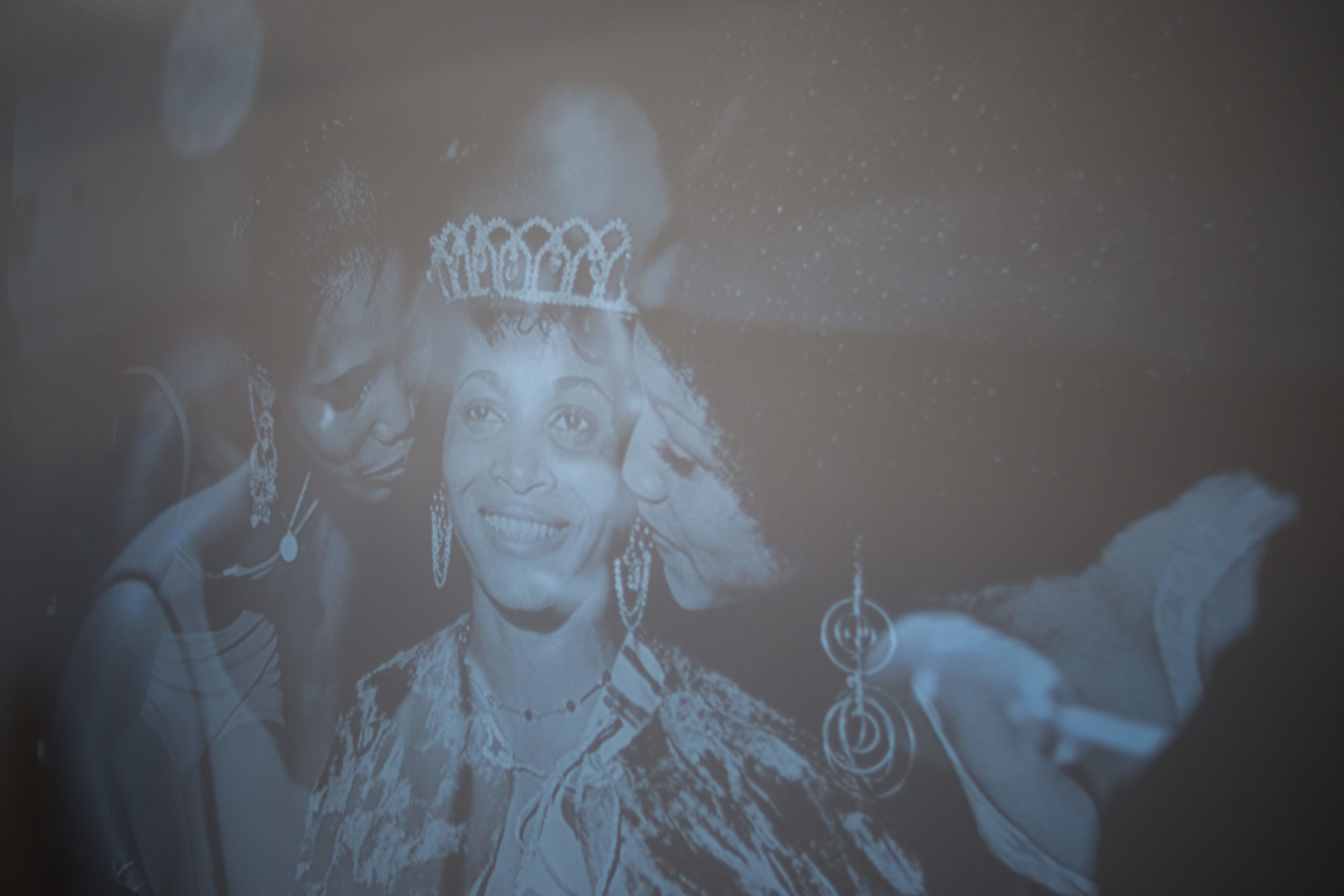 A still from a black and white video projection featuring a three people standing close together. The middle person is wearing an elaborate hairpiece and earrings. The two other people are turned towards the middle figure with eyes closed, leaning slightly inward.